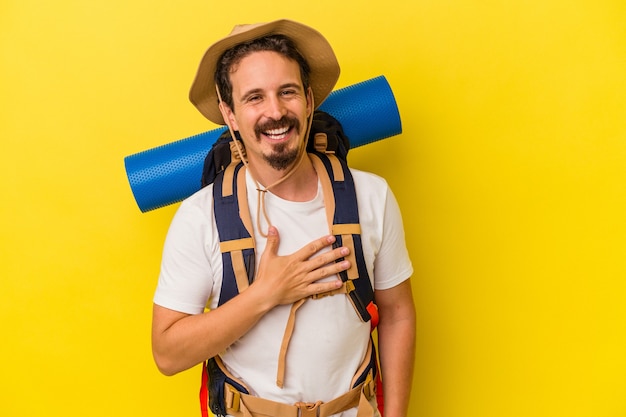 Young caucasian hiker man isolated on yellow background laughs out loudly keeping hand on chest