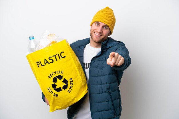 幸せな表情で正面を指している白い背景に分離されたプラスチック ボトルの完全な袋を保持している若い白人をリサイクルします。