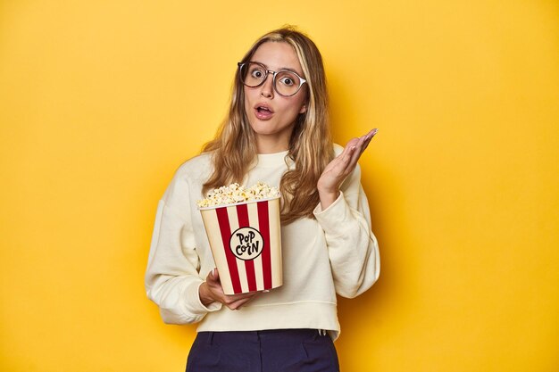 Young Caucasian holding popcorn cinema concept on yellow backdrop surprised and shocked