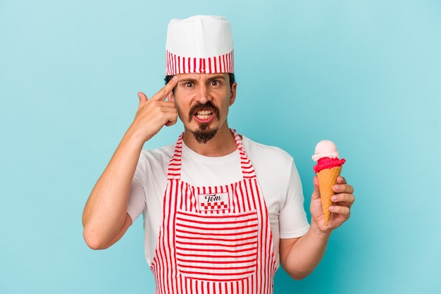 Young caucasian ice cream maker holding a ice cream isolated on blue background showing a disappointment gesture with forefinger.