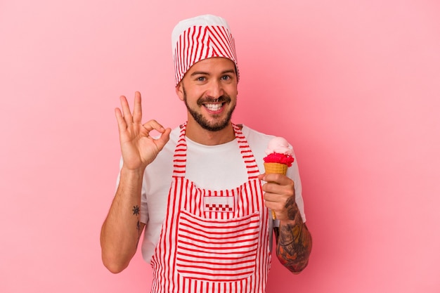 Young caucasian ice maker man with tattoos holding ice cream isolated on pink background  cheerful and confident showing ok gesture.