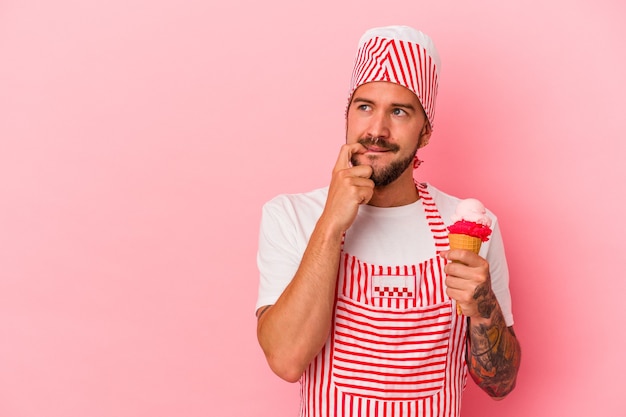 Young caucasian ice maker man with tattoos holding ice cream isolated on pink background  relaxed thinking about something looking at a copy space.