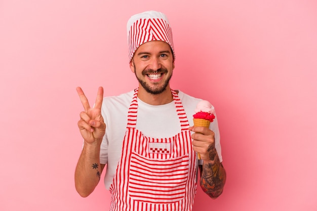 Young caucasian ice maker man with tattoos holding ice cream isolated on pink background  showing number two with fingers.