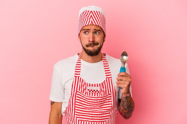 Young caucasian ice maker man with tattoos holding spoon isolated on pink background  confused, feels doubtful and unsure.