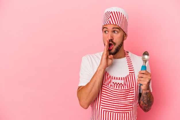 Young caucasian ice maker man with tattoos holding spoon\
isolated on pink background is saying a secret hot braking news and\
looking aside
