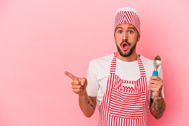 Young caucasian ice maker man with tattoos holding spoon isolated on pink background  pointing to the side