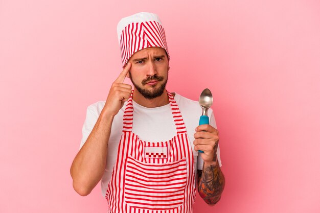 Young caucasian ice maker man with tattoos holding spoon isolated on pink background  pointing temple with finger, thinking, focused on a task.