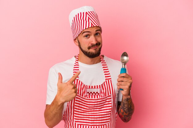 Young caucasian ice maker man with tattoos holding spoon isolated on pink background  pointing with finger at you as if inviting come closer.