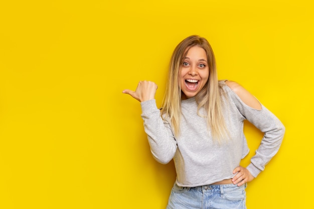 Una giovane donna bionda sorridente impressionata caucasica in un abbigliamento casual indica lo spazio vuoto vuoto per il testo o il design con il dito che presenta il prodotto isolato su un muro giallo di colore brillante