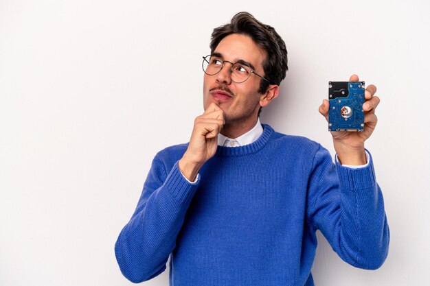 Young caucasian informatic man holding a hard disc drive isolated on white background looking sideways with doubtful and skeptical expression