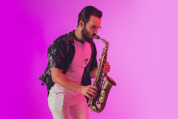 Photo young caucasian jazz musician playing the saxophone on gradient pink studio background in neon light. concept of music, hobby, festival. joyful, cheerful attractive guy. colorful portrait of artist.