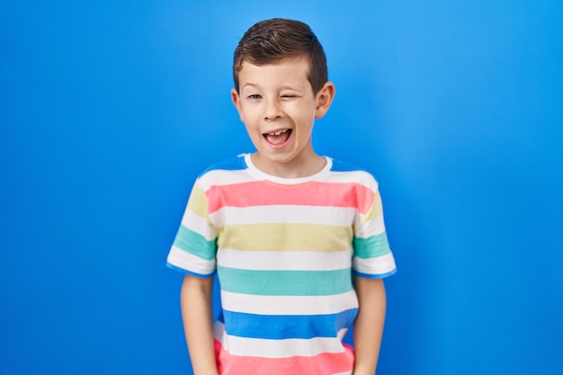 Young caucasian kid standing over blue background winking looking at the camera with sexy expression cheerful and happy face