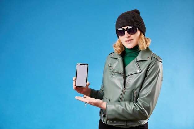 白人の若い女性は、青の背景に幸せを感じるカジュアルな服装に身を包んだ肯定的な表情の笑顔で空のスマートフォン画面を示しています女性の手で白い画面を持つ携帯電話