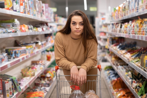 スーパーマーケットの若い白人女性カメラを見てカートに寄りかかって選択の大規模な品揃えの複雑さ買い手は食べ物を買う
