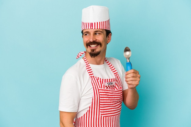Young caucasian maker holding a scoop isolated on blue background looks aside smiling, cheerful and pleasant.