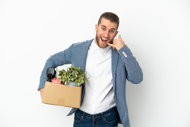 Young caucasian making a move while picking up a box full of things isolated on white wall making phone gesture. Call me back sign