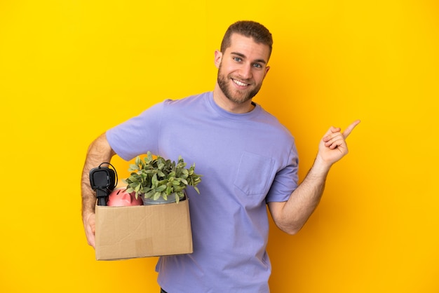 Young caucasian making a move while picking up a box full of things isolated on yellow pointing back