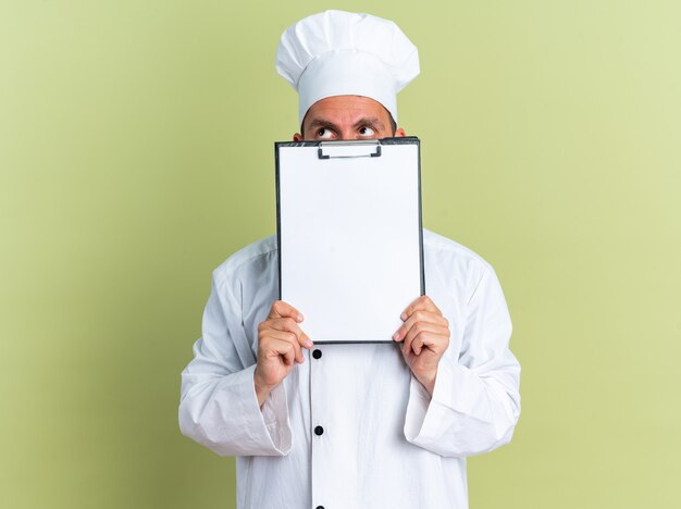 Foto giovane cuoco maschio caucasico in uniforme da chef e cappuccio che tiene appunti guardando in alto da dietro isolato su parete verde oliva