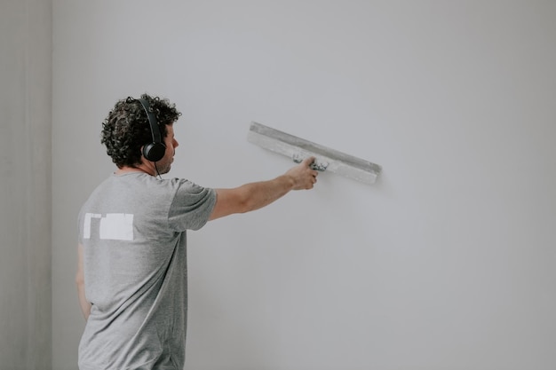 Young caucasian male plasterer listens to music through headphones and holds a large spatula on the wall