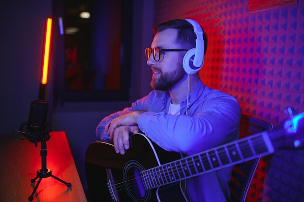 Young Caucasian male singer in headphones hold play guitar record new single on smartphone at home studio Millennial man artist use musical instrument sing shoot music video on cellphone camera