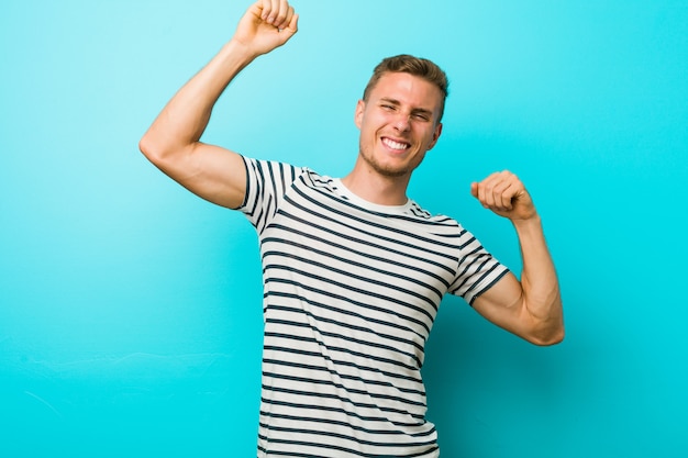 Young caucasian man against a blue wall celebrating a special day, jumps and raise arms with energy.