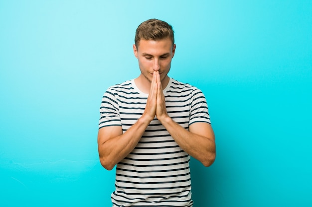 Young caucasian man against a blue wall holding hands in pray near mouth, feels confident.