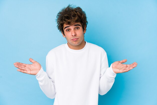 Young caucasian man against a blue wall isolated doubting and shrugging shoulders in questioning gesture.