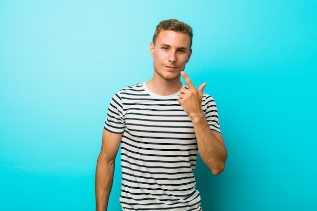 Young caucasian man against a blue wall pointing with finger at you as if inviting come closer