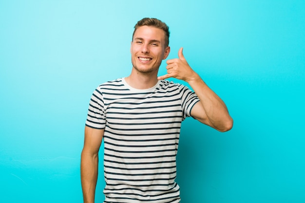 Young caucasian man against a blue wall showing a mobile phone call gesture with fingers.