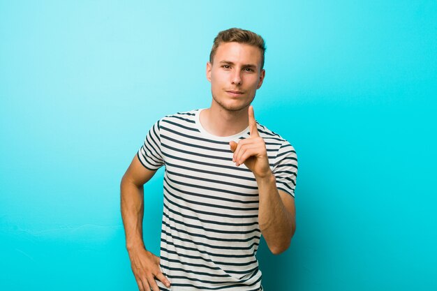 Young caucasian man against a blue wall showing number one with finger.