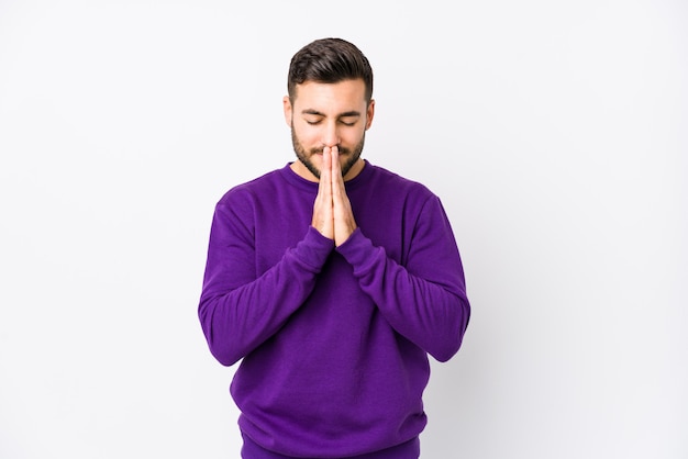 Young caucasian man against a white wall holding hands in pray near mouth, feels confident.