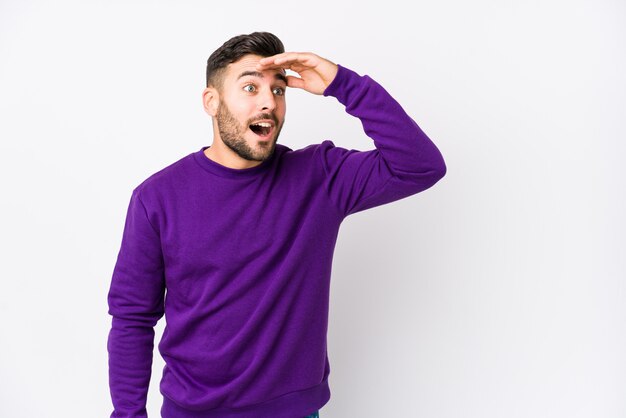 Young caucasian man against a white wall looking far away keeping hand on forehead.