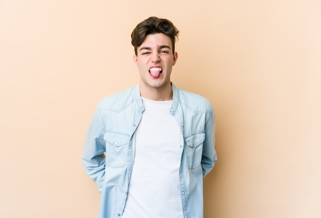 Young caucasian man on beige wall funny and friendly sticking out tongue.