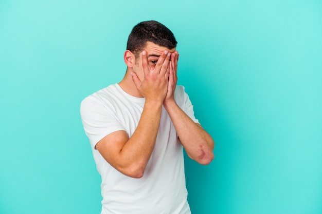 Young caucasian man on blue blink through fingers frightened and nervous.