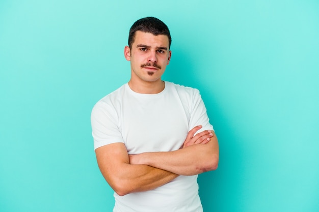 Young caucasian man on blue frowning face in displeasure, keeps arms folded.