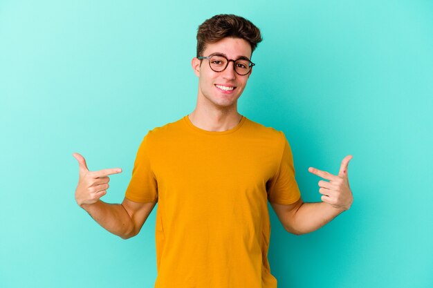 Young caucasian man on blue points down with fingers, positive feeling.