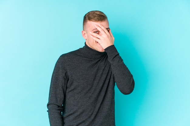 Young caucasian man on a blue wall blink at the camera through fingers, embarrassed covering face.