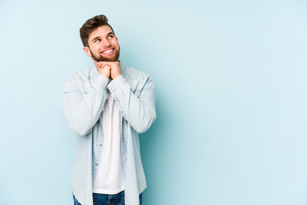 Young caucasian man on blue wall keeps hands under chin, is looking happily aside.