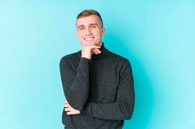 Young caucasian man on a blue wall smiling happy and confident, touching chin with hand.