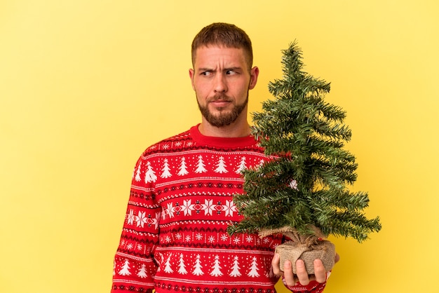 Young caucasian man buying little tree for Christmas  isolated on yellow background confused, feels doubtful and unsure.