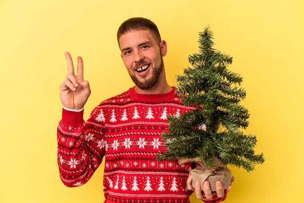指で平和のシンボルを示す黄色の背景に楽しくてのんきなクリスマスのために小さな木を購入する若い白人男性。