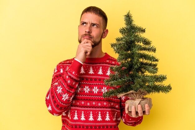 젊은 백인 남자는 의심스럽고 회의적인 표정으로 옆을 바라보며 노란색 배경에 고립된 크리스마스를 위해 작은 나무를 구입합니다.