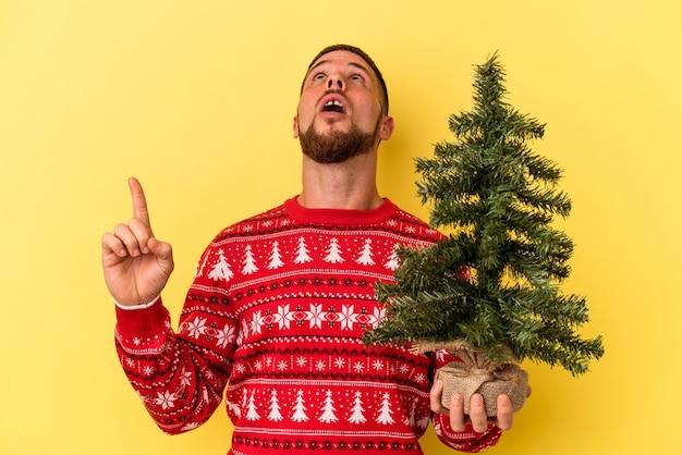 Young caucasian man buying little tree for Christmas  isolated on yellow background pointing upside with opened mouth.