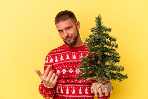 誘うようにあなたに指を指して黄色の背景に分離されたクリスマスのために小さな木を購入する若い白人男性が近づいています。