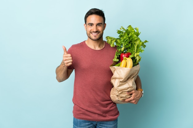 Young caucasian man buying some vegetables isolated on blue background giving a thumbs up gesture