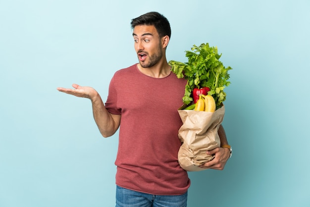 Young caucasian man buying some vegetables isolated on blue holding copyspace imaginary on the palm