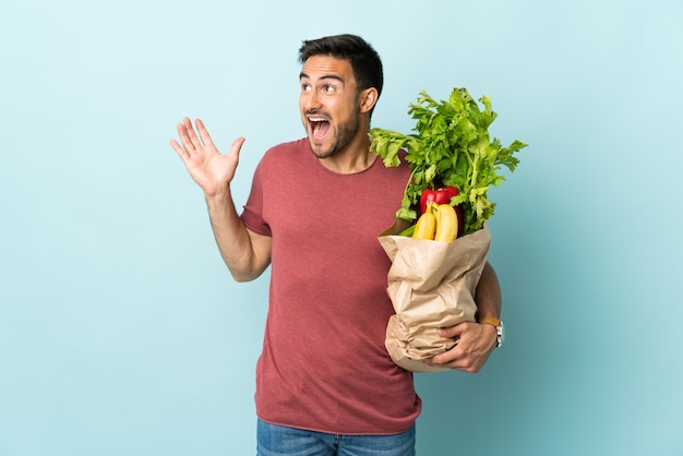 Young caucasian man buying some vegetables isolated on blue wall with surprise facial expression