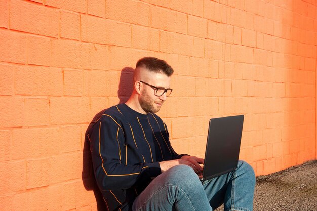 Giovane uomo caucasico in abbigliamento casual seduto a gambe incrociate per terra e al lavoro su laptop freelancer che lavora fuori digitando sul computer portatile parete rossa sullo sfondo