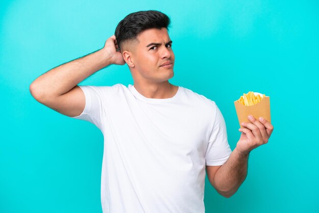 Young caucasian man catching french fries isolated on blue background having doubts