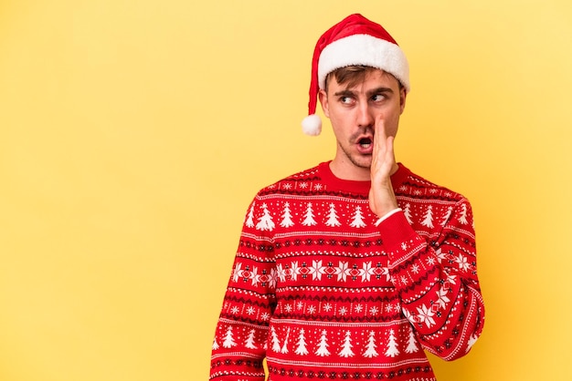 Young caucasian man celebrating Christmas isolated on yellow background is saying a secret hot braking news and looking aside
